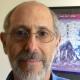 photo of bearded and balding white male with glasses, next to a poster of an erupting volcano 